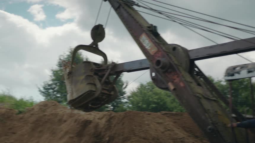 A large vintage crane with a clamshell bucket is in the process of unloading dirt or gravel onto a growing pile under a cloudy sky in an outdoor work area