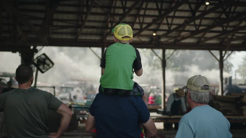 A child wearing a green shirt and yellow cap sits on an adults shoulders at a busy outdoor market under a large pavilion surrounded by people