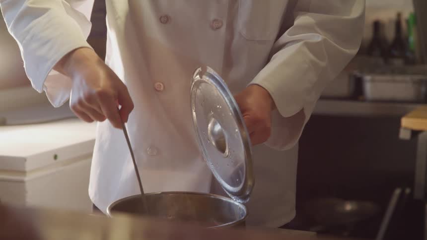 A person in a white chefs coat stirs something in a pot while holding the lid