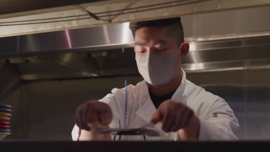 A man in a kitchen wearing a face mask and white coat takes an order and places it on a pin above the counter