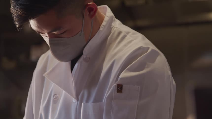 A chef wearing a white coat and face mask looks down intently while working in a kitchen