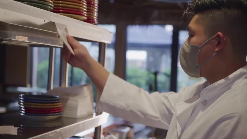 A chef in a white coat and face mask takes an order in a restaurant and secures it onto a pin
