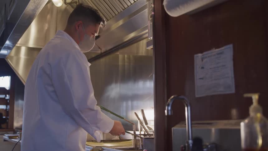 A person in a white coat shakes a fryer basket in a restaurant kitchen