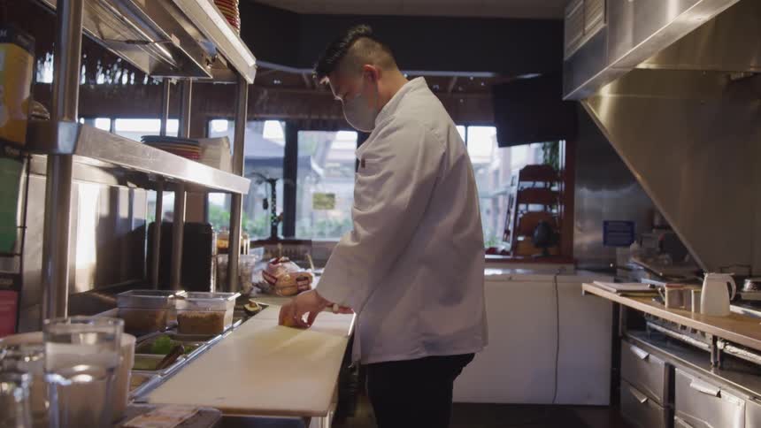 A chef in a restaurant kitchen with large windows overlooking the street cuts ingredients on a chopping board