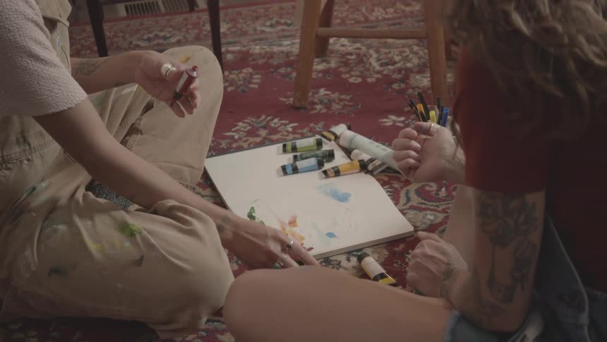 A person in brown overalls and a teenager with a tattooed arm are finger painting with oils on a notebook placed on the carpeted floor