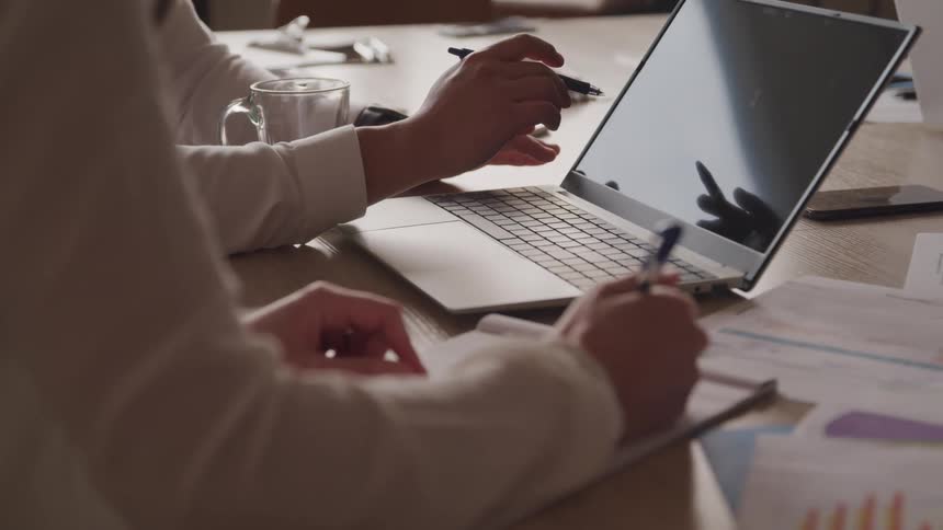Two individuals are working at a table one using a laptop and holding a pen near the screen while the other is writing on a notepad with papers and a mug also present on the table