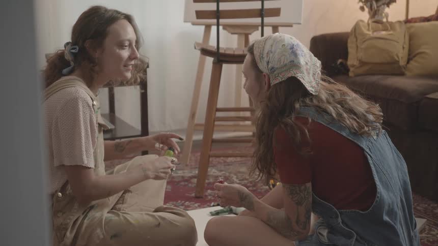 Two girlfriends sit on the floor squeezing paint from tubes and laughing