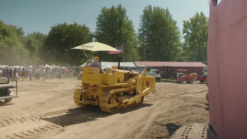 A yellow caterpillar bulldozer with an umbrella and an american flag attached drives through an outdoor event with machinery and people in the background