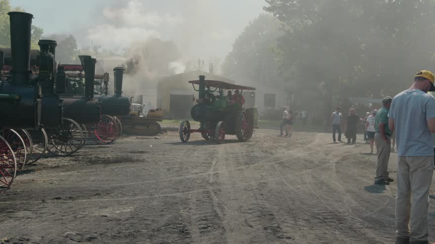 A vintage steam tractor drives along a dirt road surrounded by spectators and other historical steam engines in the background