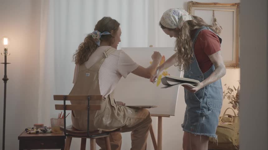 Two women are painting a yellow sunflower on a canvas indoors