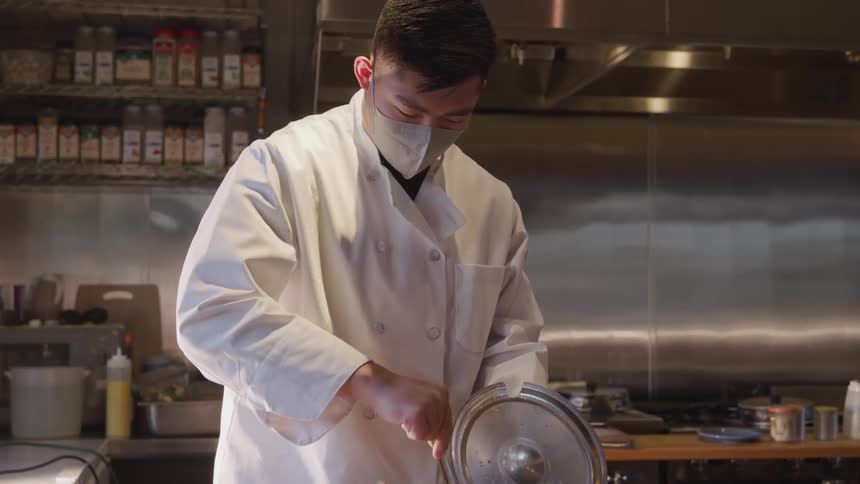 A chef wearing a face mask stirs a pot in a restaurant kitchen