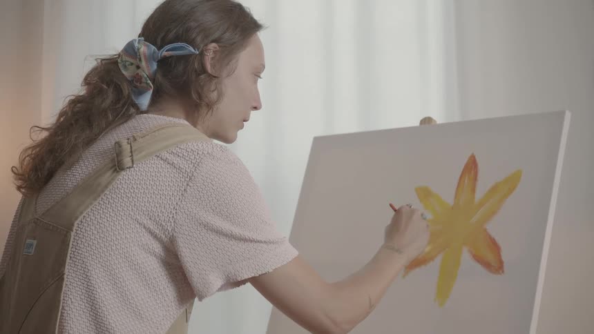 A closeup of a woman wearing brown overalls painting a flower on a canvas set on an easel using a brush in a welllit room