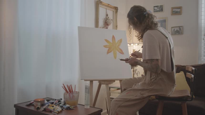 A young artist with curly hair wearing overalls is seated and painting a yellow flower on a white canvas propped on an easel in a room with soft natural light and various art supplies nearby