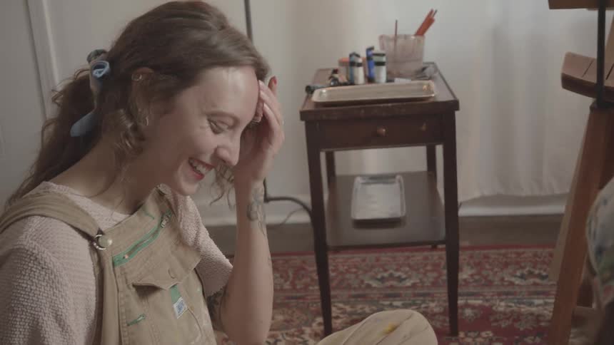 A woman in a beige overall sits on a carpeted floor in a room painting her fingers alongside a friend