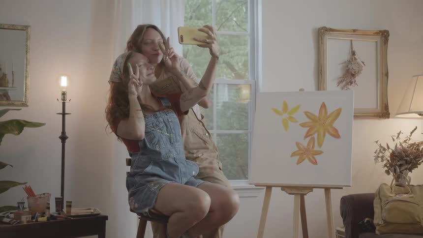 Two young adults are taking a selfie with a smartphone in a brightly lit room with one sitting on a stool and the other standing behind near a painting of yellow flowers on an easel and various household items