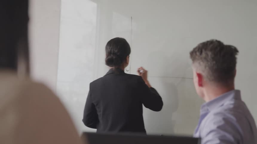 A person writes on a whiteboard while another watches attentively in a well lit office setting