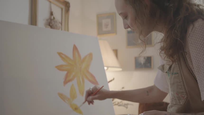 A close up of a woman artist in a beige overall painting flowers on a canvas at an easel in a cozy room