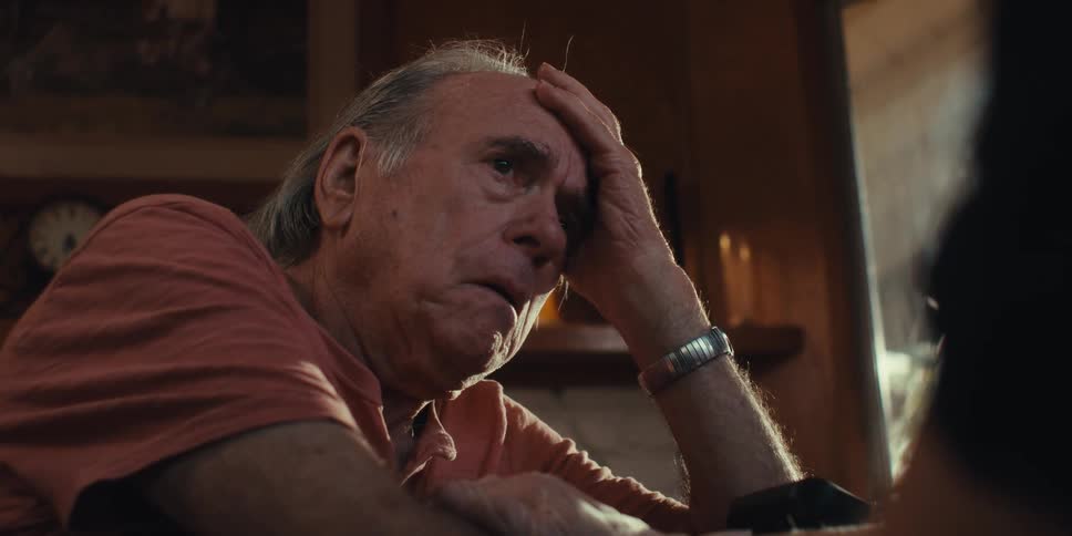 An elderly man sits at home crying while another person holds his hands to console him