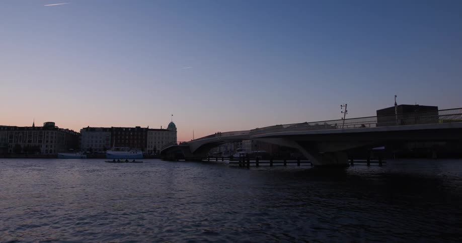 A kayak rowing team paddles under a bridge near a city during twilight with a blue sky overhead
