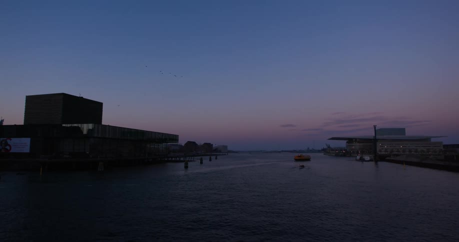 A modern waterfront at dusk with buildings flanking a calm waterway and a single boat drifting under the clear gradient sky
