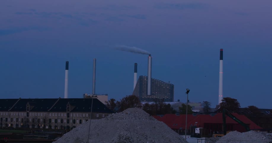 An industrial area at dusk features tall chimneys emitting smoke a large gravel pile in the foreground and a mix of buildings in the background