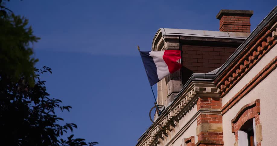 The french flag waves from a pole attached to the side of a lightcolored building with brick accents under a clear blue sky