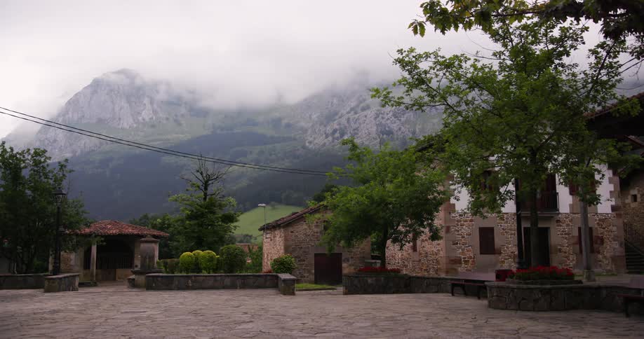 A quaint village square surrounded by stone buildings vibrant gardens and rolling hills under a misty mountain backdrop