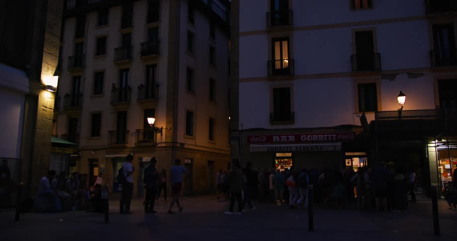 A dimly lit street scene at dusk shows groups of people gathered outside buildings