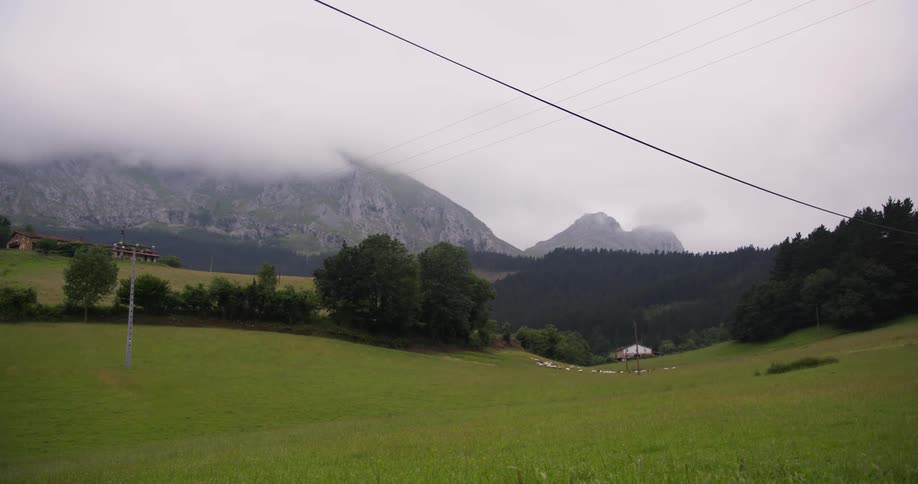 Slowmoving clouds drift over misty mountain peaks as a distant herd of sheep grazes quietly on a wide green pasture near a small farmhouse