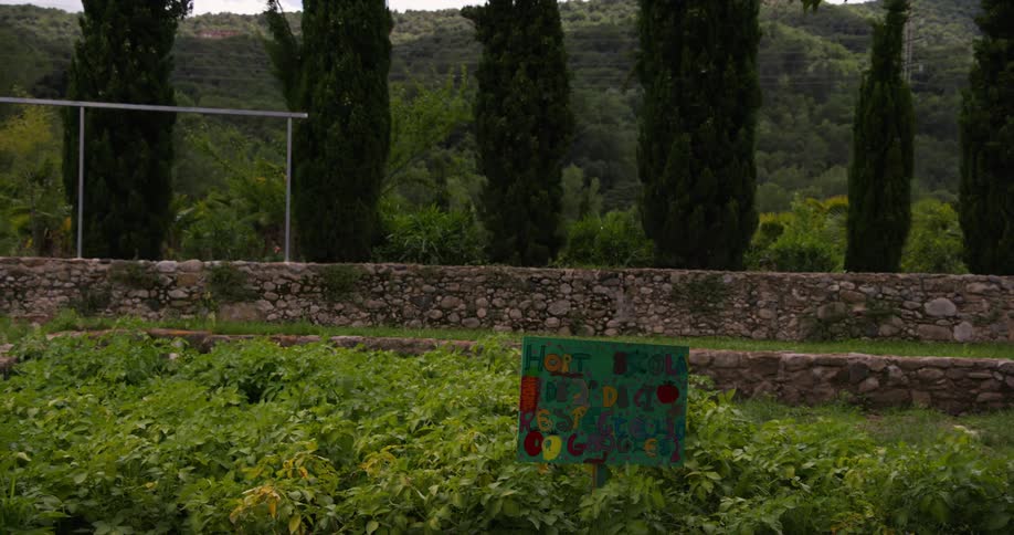 A lush garden features a colorful sign surrounded by stone walls tall cypress trees and rolling green hills in the background