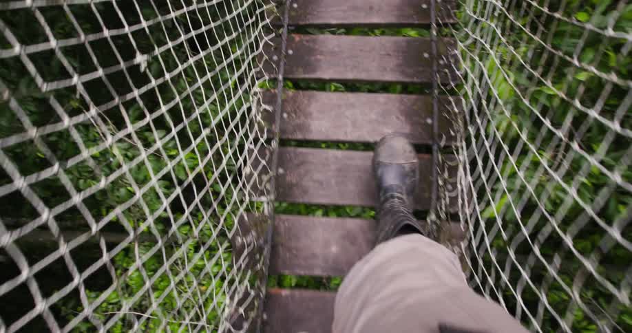 A person walks across a wooden suspension bridge with netted sides surrounded by dense green foliage below