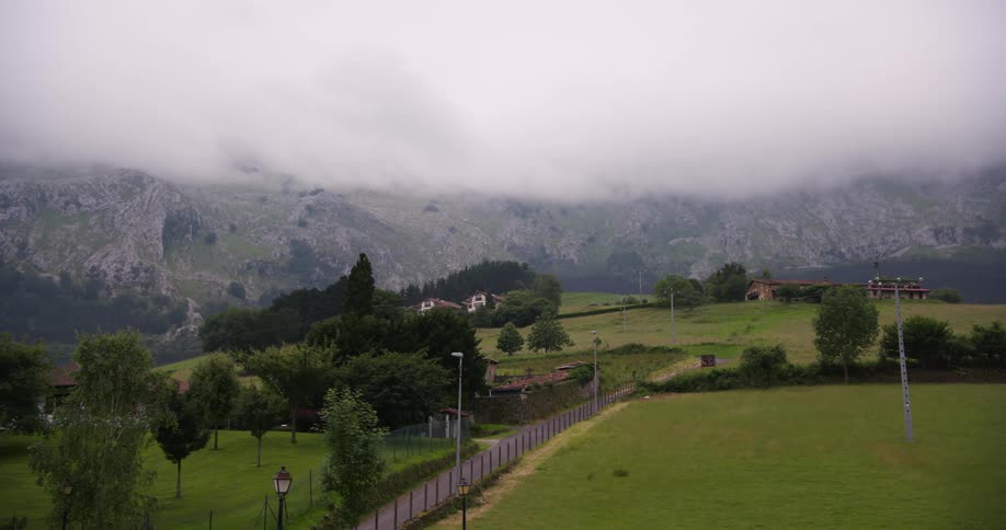 Clouds drift slowly across the mountain tops above a quiet stretch of countryside dotted with scattered homes green pastures and winding rural paths