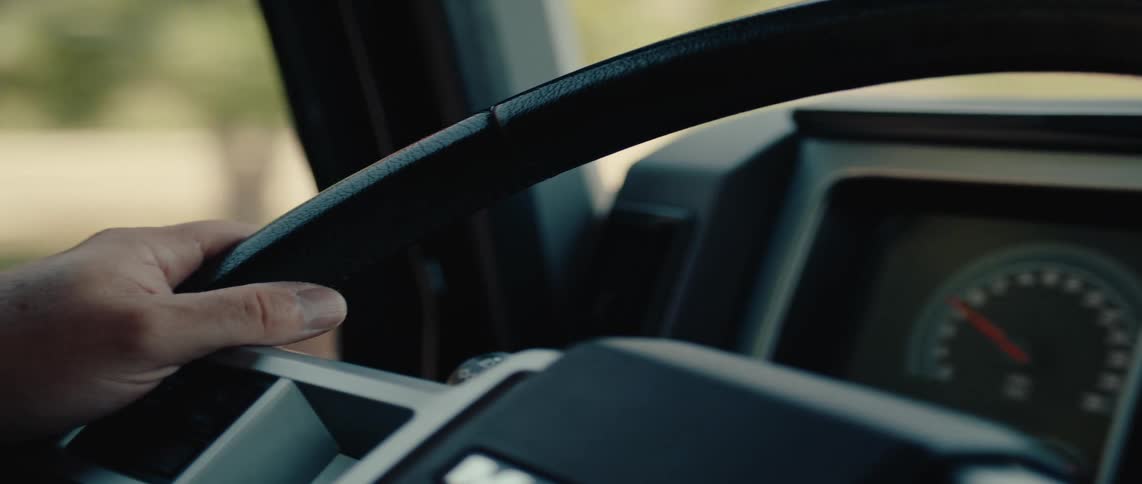 A hand gripping the steering wheel of a vehicle with a blurred view of the dashboard and the outside visible through the windshield