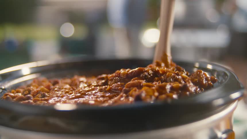 A hand stirs a pot of chili with a wooden spoon at an outdoor gathering