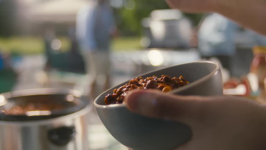 A hand serving chili with a wooden spoon at an outdoor gathering