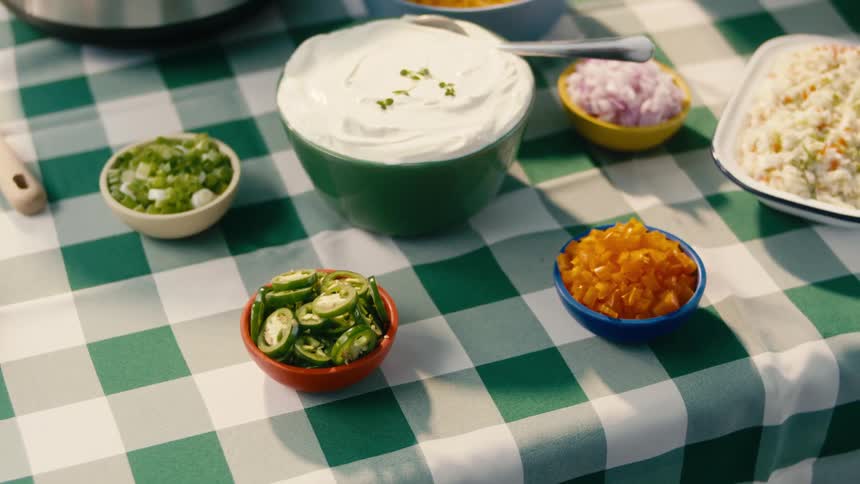 Small bowls of garnish next to large dip and potato salad on a checkered tablecloth