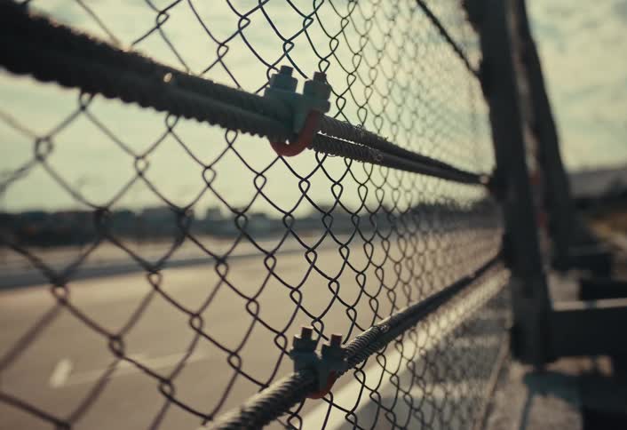 A closeup shot shows a chainlink fence secured with metal fasteners to horizontal bars with a blurred background suggesting an outdoor urban or industrial area