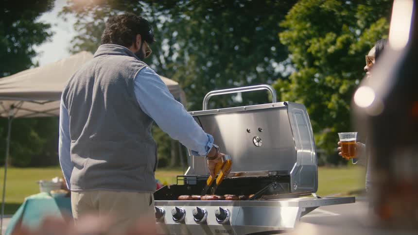 A person grills hot dogs on a stainless steel barbecue talking to a woman holding a drink