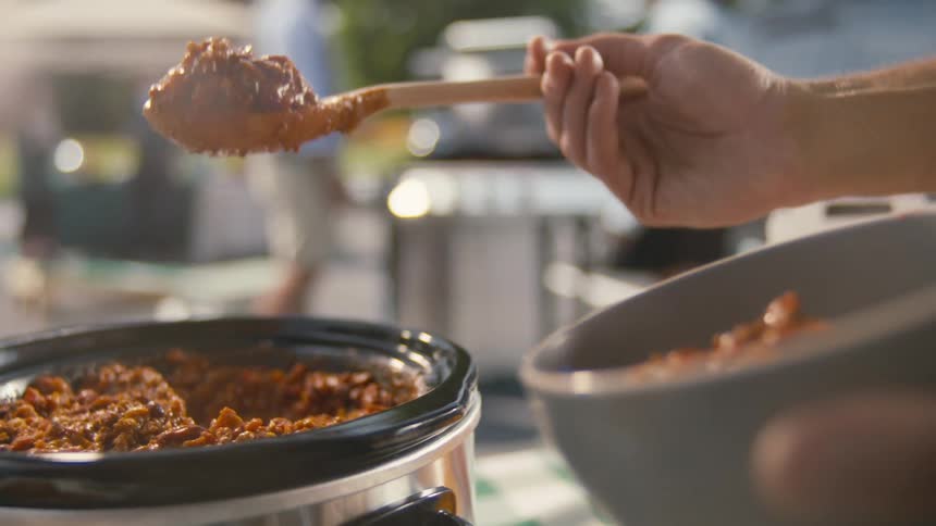 A person serves a spoonful of chili into a bowl during an outdoor bbq
