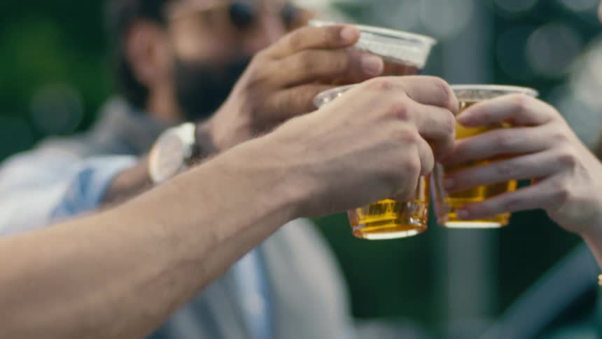 A closeup shows a group of friends raising plastic cups filled with beer while toasting outdoors