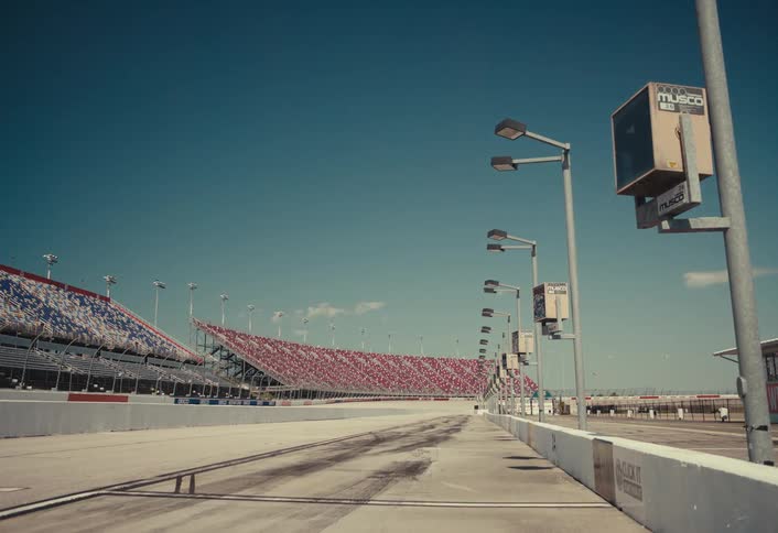 An empty pit lane stretches beneath stadium lighting beside colorful grandstands in a wide open speedway as tire marks and sponsor boards line the concrete under a clear blue sky
