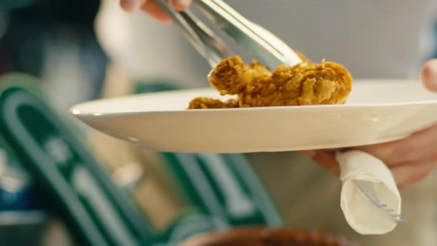 A hand uses metal tongs to lift a piece of crispy fried chicken from a plate piled with other pieces of fried chicken