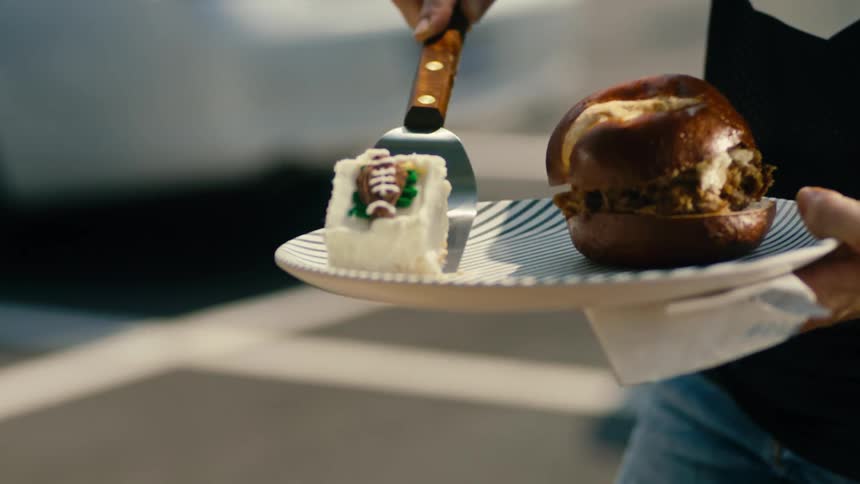 A person serves a slice of cake on a plate with a hamburger at an outdoors bbq