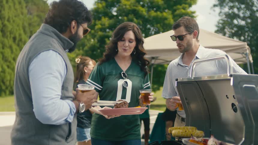 People gather around a barbecue grill holding drinks and plates with corn on the cob and various grilled food visible on the grill