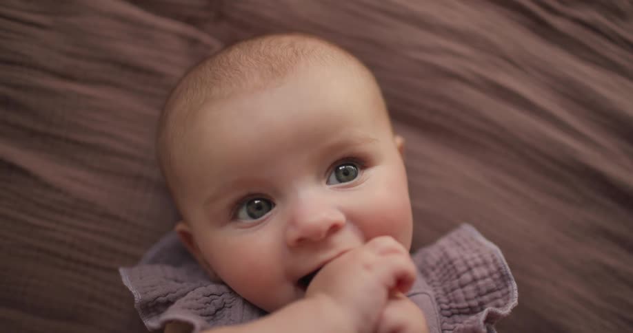A baby with light blue eyes and fair skin lies on a textured brown surface looking up with a hand near their mouth