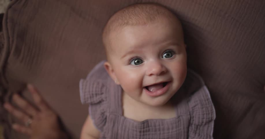 A happy baby with bright eyes and a slightly open mouth wearing a ribbed purple outfit and lying against a brown textured surface with a glimpse of an adults hand nearby