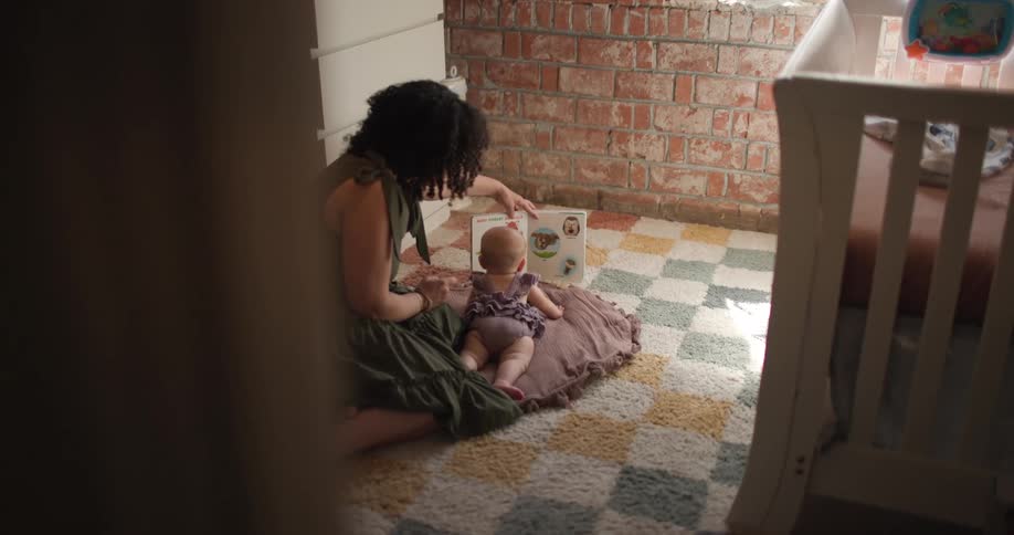 A mother is sitting on the floor reading a picture book to a baby lying on a blanket in a cozy sunlit nursery