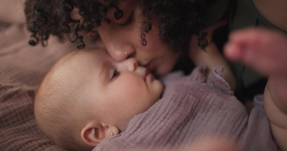 A baby laying in bed is getting kissed by a woman presumably its mother