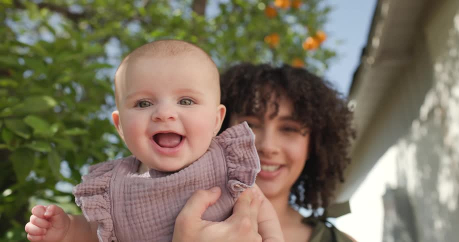 The face of a little baby who is very happy and is in the lap of a woman and they are in an outdoor environment and the baby is wearing the pink dress