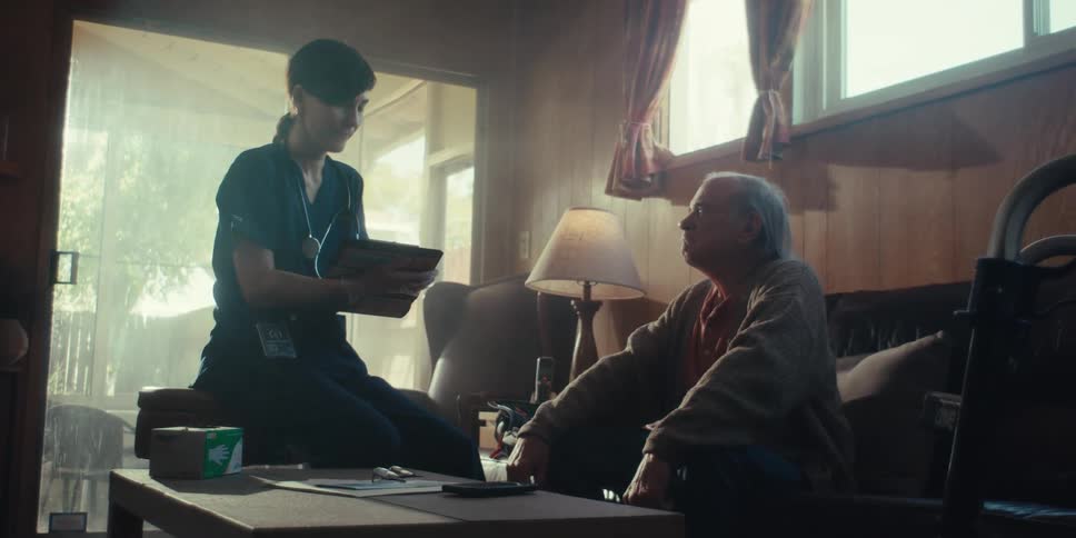 A healthcare worker in dark scrubs kneels beside an elderly person seated in a chair reviewing information on a clipboard